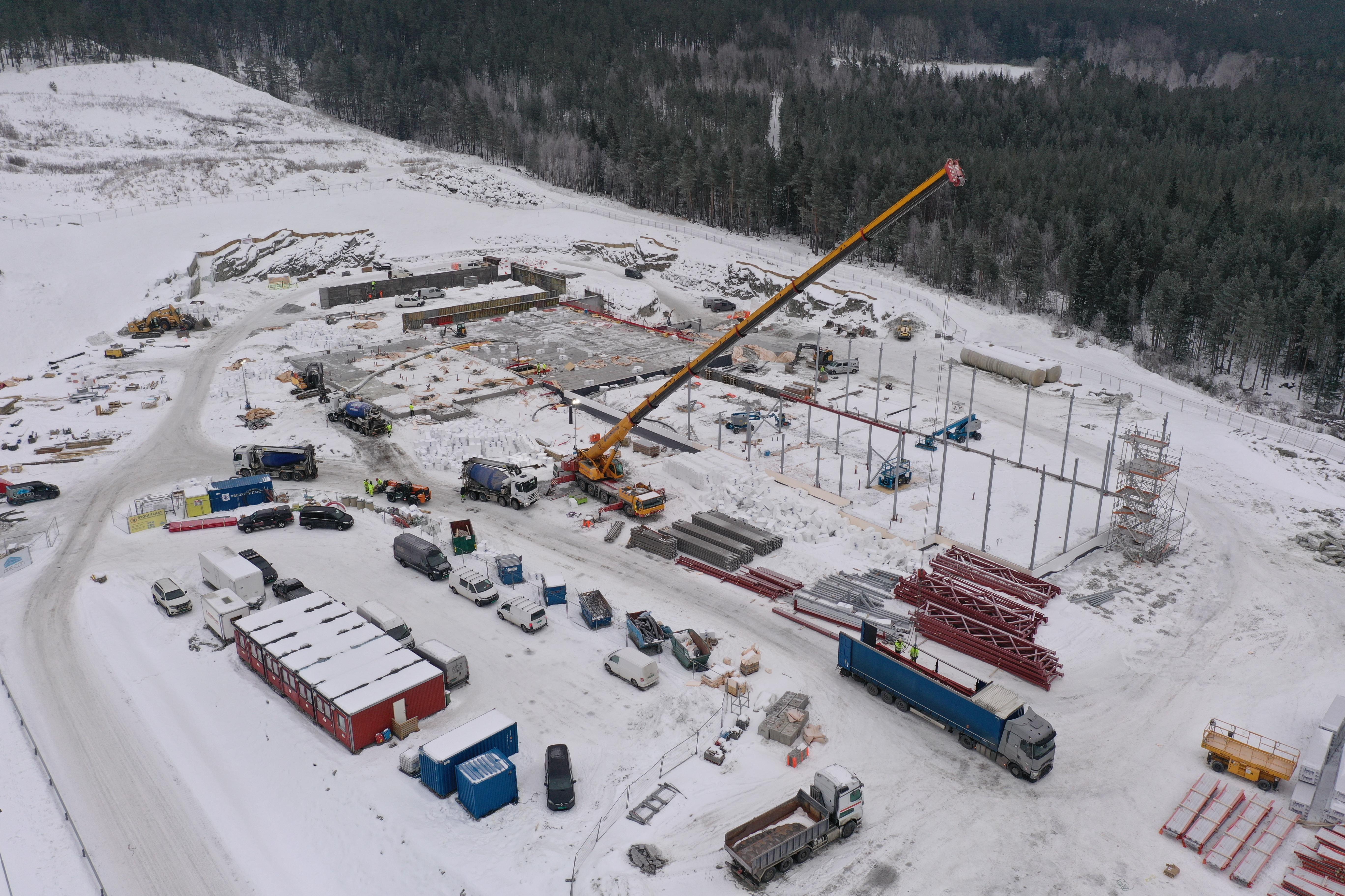 Stor byggeplass i snøen sett fra lufta, med kran, stålramme og brakker